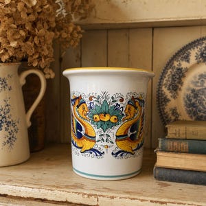 May include: A white ceramic cylindrical utensil holder featuring a hand-painted design of two yellow and blue mythical sea creatures facing a central green and orange floral motif, finished with a solid yellow rim and a thin blue base stripe.