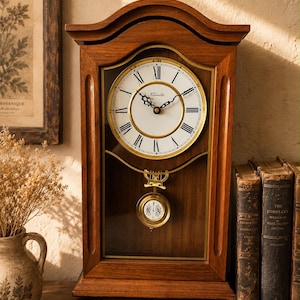 Puede incluir: Un reloj de sobremesa de madera con marco rectangular y parte superior arqueada. La esfera es blanca con números romanos negros y el texto Forestville. Un péndulo dorado con las letras RA cuelga tras un panel de cristal central.