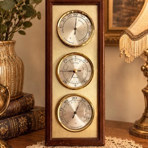 May include: A rectangular weather station featuring a dark wood frame and a brushed gold-tone metal face. It contains three circular analogue gauges: a hygrometer at the top, a barometer in the centre, and a thermometer at the bottom with Celsius.