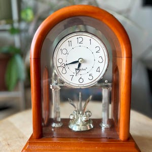 May include: A Westclox mantel clock with a wooden arched frame and a glass front. The white clock face features black Arabic numerals and ornate hands. Below the clock face, a silver-colored pendulum with four spheres rotates between two silver pillars.