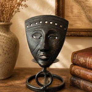 Puede incluir: Una escultura de metal negro que presenta una máscara de rostro humano estilizada montada sobre una base circular. La máscara tiene una superficie texturizada, una fila de pequeños recortes circulares en el borde superior y un soporte.