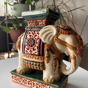 Peut inclure: Un tabouret ou support de plante en céramique en forme d'éléphant, au corps crème orné de motifs décoratifs orange, bleus et verts. L'objet présente un plateau et une base rectangulaires avec des sculptures en relief détaillées et une finition brillante.