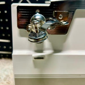 May include: A white electric can opener with a metallic cutting mechanism and a magnetic lid lifter. The device features a horizontal gray stripe near the base and the brand name RIVAL printed in white lettering on the lower front section.