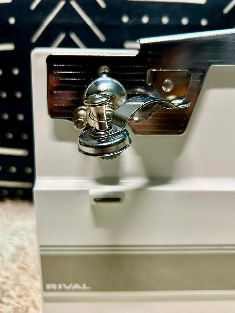 May include: A white electric can opener with a metallic cutting mechanism and a magnetic lid lifter. The device features a horizontal gray stripe near the base and the brand name RIVAL printed in white lettering on the lower front section.