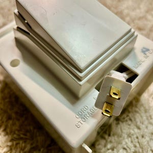 May include: A white plastic cord storage device featuring a large rocker switch on top. The unit has a rectangular base with the text CORD STORAGE embossed on the side and a two-prong electrical plug extending from a recessed compartment.