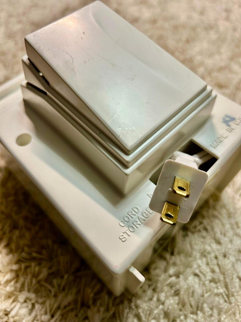 May include: A white plastic cord storage device featuring a large rocker switch on top. The unit has a rectangular base with the text CORD STORAGE embossed on the side and a two-prong electrical plug extending from a recessed compartment.