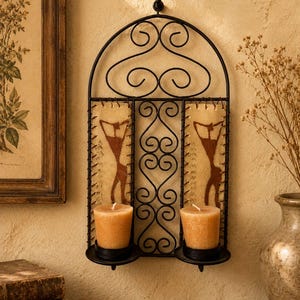 May include: A black metal wall-mounted candle holder with an arched top and scrollwork design. It features two rectangular panels with brown abstract figure illustrations and two circular platforms holding light orange votive candles.