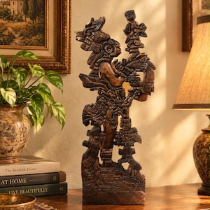 May include: A dark brown, intricately carved wooden sculpture depicting a figure in elaborate ceremonial attire and headdress, standing in profile while holding a tall, ornate staff. The carving features detailed textures and relief patterns.
