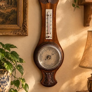 May include: A wall-mounted weather station featuring a dark wood frame with carved floral accents at the top and bottom. It includes a vertical thermometer with Fahrenheit and Celsius scales and a circular barometer with a silver dial and text.