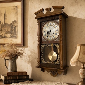 Puede incluir: Un reloj de pared rectangular de madera con acabado marrón oscuro, que presenta una esfera circular con números romanos negros, detalles dorados en las esquinas y un péndulo color latón visible tras un panel de cristal con filigrana dorada.
