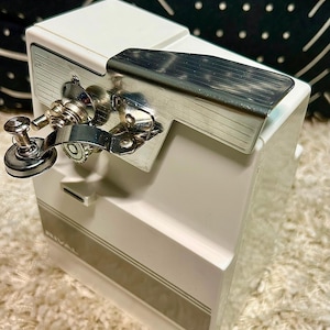 May include: A white electric can opener with a chrome-plated metal lever and cutting mechanism. The device features a rectangular body with a gray horizontal stripe and the brand name RIVAL printed in white on the lower front section.