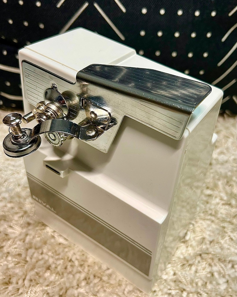 May include: A white electric can opener with a chrome-plated metal lever and cutting mechanism. The device features a rectangular body with a gray horizontal stripe and the brand name RIVAL printed in white on the lower front section.