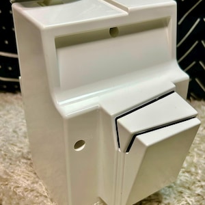 May include: A white plastic wall-mounted cord storage unit featuring a rectangular body with a hinged top lid, a protruding front compartment, and a small circular hole on the side. The words CORD STORAGE are embossed on the lower front section.