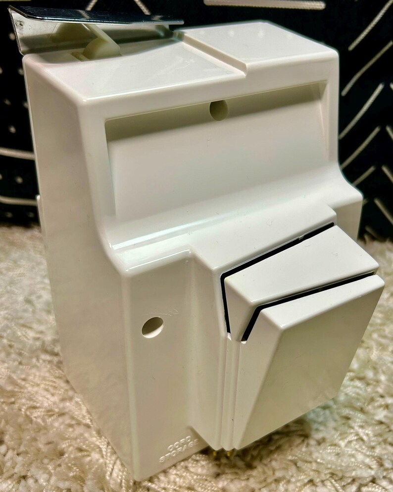 May include: A white plastic wall-mounted cord storage unit featuring a rectangular body with a hinged top lid, a protruding front compartment, and a small circular hole on the side. The words CORD STORAGE are embossed on the lower front section.