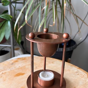 May include: A copper-colored metal oil burner featuring a circular base and a top platform supported by three vertical pillars. The top platform has a central bowl for oil and three small spherical accents. A small white tea light candle sits below.