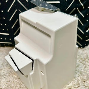 May include: A white plastic wall-mounted dispenser featuring a rectangular body with a slanted front compartment and a metallic silver lever at the top. The device has a smooth finish and a functional design with a small circular opening on the side.