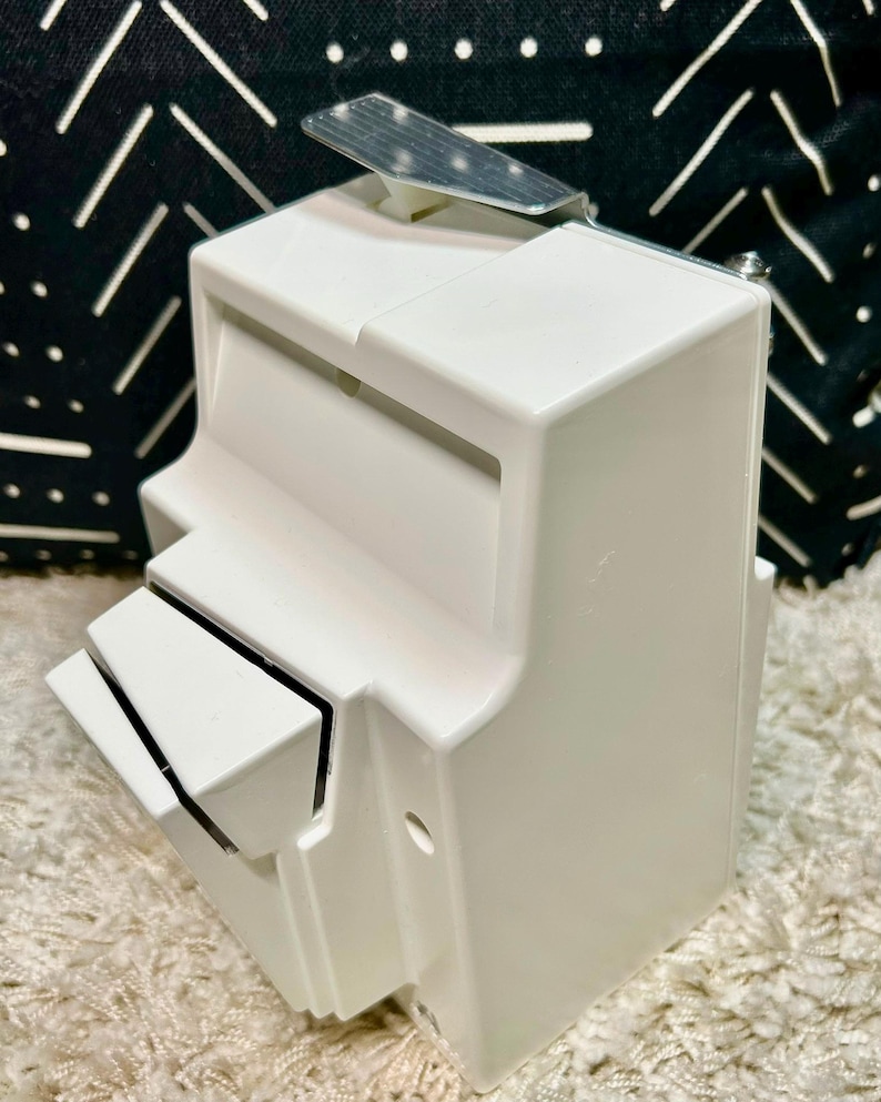 May include: A white plastic wall-mounted dispenser featuring a rectangular body with a slanted front compartment and a metallic silver lever at the top. The device has a smooth finish and a functional design with a small circular opening on the side.