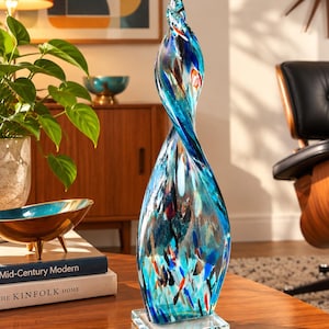 May include: A tall, hand-blown glass sculpture featuring a twisted, spiral design. The glass displays a vibrant mix of blue, turquoise, and clear tones, accented with scattered flecks of red, black, and white. It stands on a clear square glass base.