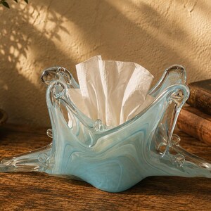 May include: A decorative tissue holder crafted from hand-blown glass featuring a swirling pattern of light blue and white colours. The glass container has an irregular, organic shape with multiple pointed edges extending outward from the centre.