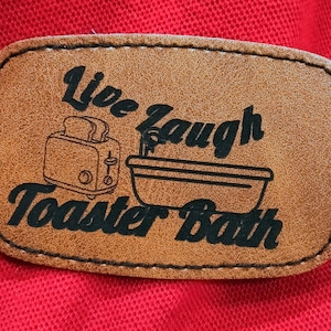 May include: A rectangular tan faux leather patch with black stitched edges. The patch features a black graphic of a toaster and a bathtub, with the black text "Live Laugh Toaster Bath" printed across the center of the surface.