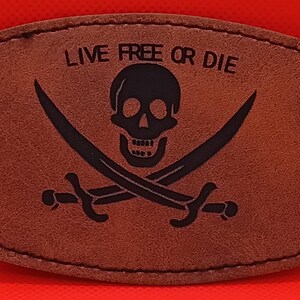 May include: A rectangular brown patch with rounded corners featuring a black graphic of a skull and two crossed swords. The text "LIVE FREE OR DIE" is printed in black above the skull. The patch has visible stitching along the outer edge.