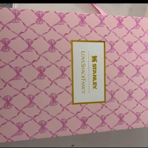 May include: A rectangular cardboard box with a light pink background featuring a repeating pattern of dark pink ribbons tied in bows. Centered on the box is a white rectangular label with a gold border, displaying the text STANLEY and LOVESHACKFANCY.