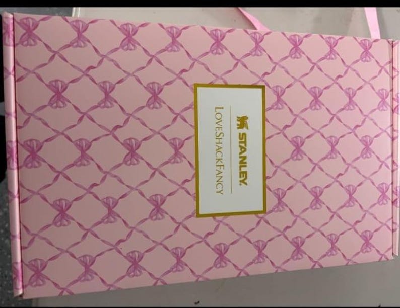 May include: A rectangular cardboard box with a light pink background featuring a repeating pattern of dark pink ribbons tied in bows. Centered on the box is a white rectangular label with a gold border, displaying the text STANLEY and LOVESHACKFANCY.