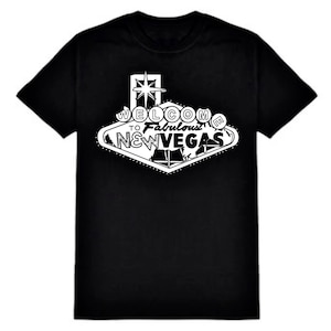 May include: A black short-sleeve t-shirt featuring a white graphic print on the chest. The design depicts a diamond-shaped sign with a star at the top and the text WELCOME TO FABULOUS NEW VEGAS, styled after a classic Las Vegas landmark sign.