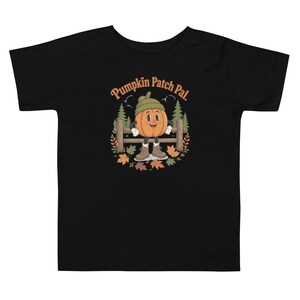May include: A black short-sleeve t-shirt featuring a graphic of a smiling pumpkin character wearing a green beanie and brown boots. The pumpkin stands before a wooden fence and pine trees, surrounded by autumn leaves, with the text Pumpkin Patch Pal.