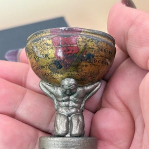 Puede incluir: Una pequeña figura metálica de tono plateado de una persona arrodillada que sostiene un globo terráqueo en forma de cuenco. El globo presenta un mapa de estilo vintage con tonos dorados y rojos, mostrando claramente South Atlantic Ocean e Indian Ocean.