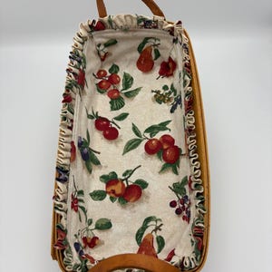May include: A rectangular woven basket with a light brown finish and two arched handles. The interior is lined with a cream-colored fabric featuring a repeating pattern of red apples, pears, and dark berries with green leaves and ruffled edges.