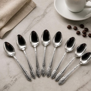 Puede incluir: Un juego de ocho cucharas de metal plateado dispuestas en abanico. Cada cuchara presenta un cuenco ovalado pulido y un mango decorativo con un patrón floral o de hojas en relieve en la base. Las cucharas tienen un acabado metálico brillante.