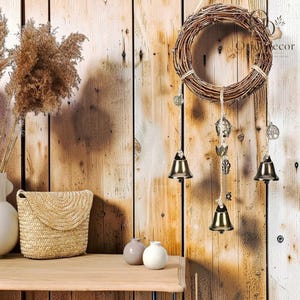 May include: A circular wreath made of brown woven twigs featuring three hanging strands of twine. The strands are adorned with small metallic bells, a pentagram charm, a tree of life charm, and a key charm. The wreath is suspended against a wall.