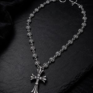 May include: A silver-toned necklace featuring a series of small, circular cross-shaped links connected by metal rings. The necklace includes a prominent, elongated cross pendant with flared ends and a toggle clasp closure at the top of the chain.
