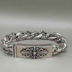 May include: A polished silver chain bracelet featuring a rectangular ID plate as the central focal point. The plate is engraved with a symmetrical floral cross motif. The bracelet is constructed from thick, interlocking curb-style links.