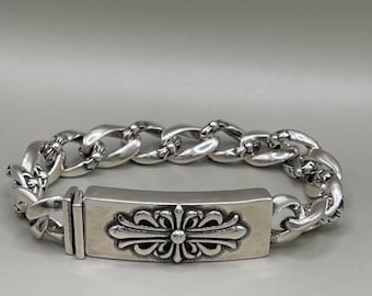 Puede incluir: Una pulsera de cadena de plata pulida con una placa de identificación rectangular como punto focal central. La placa está grabada con un emblema de cruz floral detallado. La pulsera está construida con eslabones metálicos gruesos y redondeados.