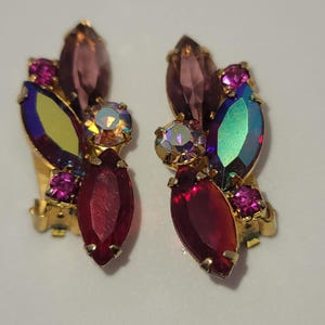 May include: A pair of gold-tone clip-on earrings featuring a cluster of multi-colored faceted rhinestones. The design includes marquise-cut stones in deep red, purple, and iridescent green, accented by small round pink stones and a central iridescent crystal.