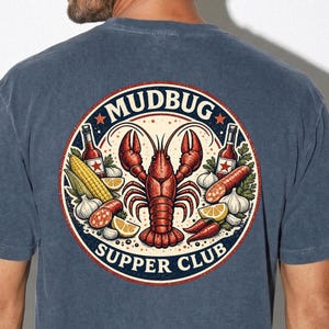 Peut inclure: Un t-shirt bleu ardoise avec un motif circulaire au dos. Le dessin montre un homard rouge au centre entouré de maïs, de saucisse, d'ail, de quartiers de citron et de bouteilles de sauce piquante. Le texte MUDBUG SUPPER CLUB entoure le bord.