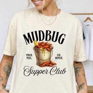 Peut inclure: Un t-shirt couleur crème avec un motif représentant un seau en métal rempli d'écrevisses orange et d'un épi de maïs. Le texte indique MUDBUG, PINCH. PEEL. EAT. REPEAT. et Supper Club en lettres noires. Le motif est centré sur le devant du t-shirt.