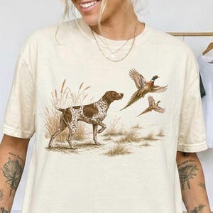 May include: A cream-colored short-sleeve t-shirt featuring a graphic illustration of a brown and white spotted hunting dog standing in tall grass, with two pheasants in flight above the dog. The design uses natural earth tones throughout.