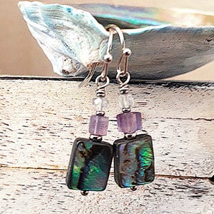 Könnte beinhalten: Ein Paar Ohrhänger mit rechteckigen Abalone-Muschel-Anhängern in schillernden Grün- und Schwarztönen. Jeder Ohrring enthält eine kleine quadratische violette Perle und eine klare facettierte Perle, die an silberfarbenen Metallhaken hängen.