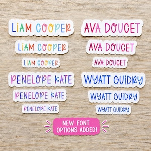 May include: A collection of custom name labels in various sizes and colors. The labels feature the names Liam Cooper in multi-colored letters, Ava Doucet in pink tones, Penelope Kate in purple, and Wyatt Guidry in blue. A pink sign reads NEW FONT OPTIONS ADDED!