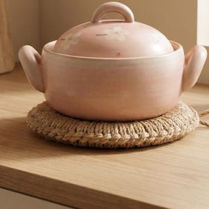 Pode incluir: Uma panela redonda de cerâmica rosa claro com duas alças laterais e uma tampa a condizer com um padrão floral branco. A panela assenta num descanso circular de juta entrançada, apresentando um acabamento vidrado e liso.