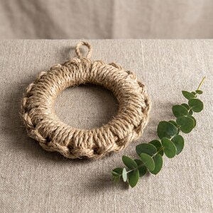 May include: A circular trivet crafted from thick, natural tan jute rope, featuring a braided texture and a small loop at the top for hanging. Beside the round rope trivet lies a single sprig of green eucalyptus leaves with small, rounded foliage.