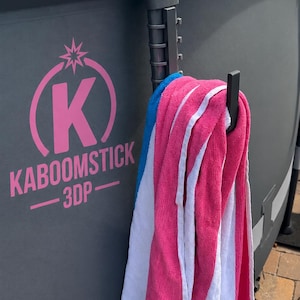 May include: A black plastic hook mounted on the side of a gray above-ground pool wall. The hook holds a striped towel in shades of pink, white, and blue. The pool wall features a pink logo with a star, the letter K, and the text KABOOMSTICK 3DP.