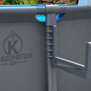 May include: A gray plastic pool accessory hook attached to the vertical frame of a gray above-ground pool wall. The pool wall features the printed text KABOOMSTICK 3DP with a star icon above the letter K. The hook has a multi-slotted adjustment design.