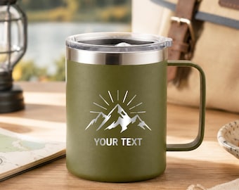Può includere: Una tazza in acciaio inossidabile verde oliva con coperchio trasparente e manico. La parte anteriore presenta una grafica argentata di una catena montuosa con raggi solari sopra e il testo "YOUR TEXT" in un carattere sans-serif sotto il disegno.