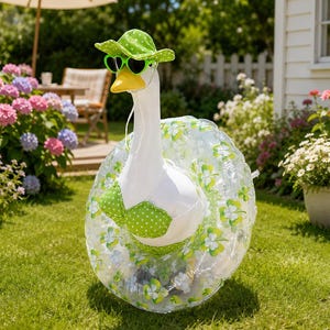 May include: A white goose figurine wearing a green polka dot sun hat, heart-shaped green sunglasses, and a green polka dot bikini top. The goose is positioned inside a clear inflatable swim ring decorated with a green and white clover pattern.