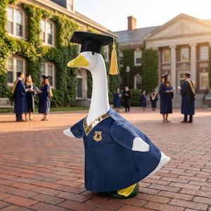 May include: A white goose figurine wearing a navy blue graduation gown with gold trim and a black mortarboard cap with a yellow tassel. The gown features a small gold emblem on the chest. The goose has a yellow beak and stands on yellow feet.