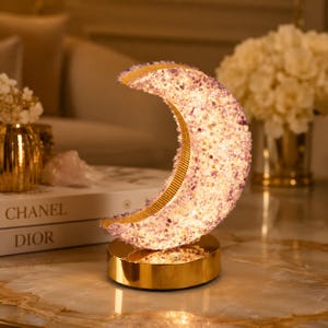 May include: A crescent moon-shaped lamp covered in small, textured purple amethyst crystals. The lamp features a gold-coloured metallic inner rim and sits on a circular gold-coloured base. It emits a warm, soft light from within the crystal structure.